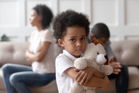 Child with a concerned expression holding a teddy bear with parents upset at each other in background